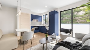 Una sala moderna con sofá, una mesa de comedor y sillas. Hay una zona de cocina con armarios azules y encimera blanca. Un ventanal ofrece vistas a árboles.