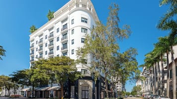 Un hotel de varios pisos con balcones, una entrada cubierta y vista a la calle con coches estacionados y árboles.
