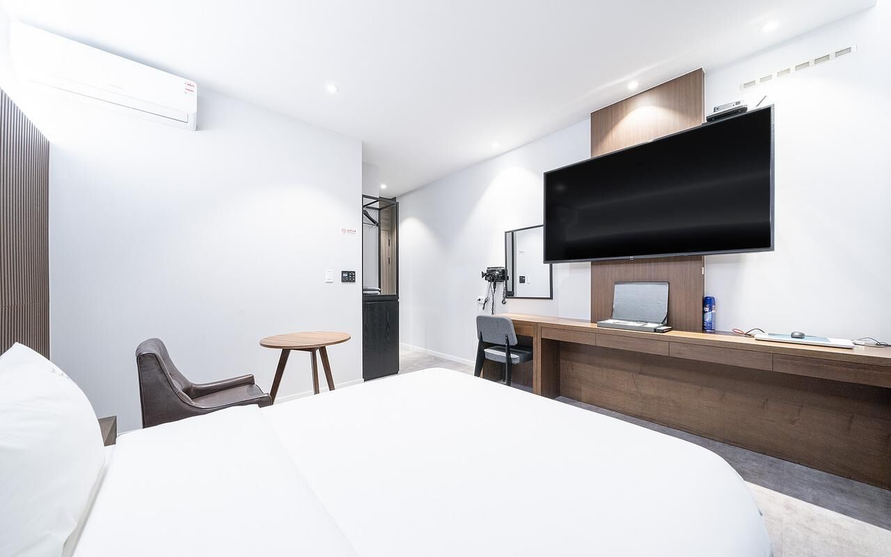 A hotel room with a bed, a desk with a computer, a chair, a small table, and a TV mounted on the wall.