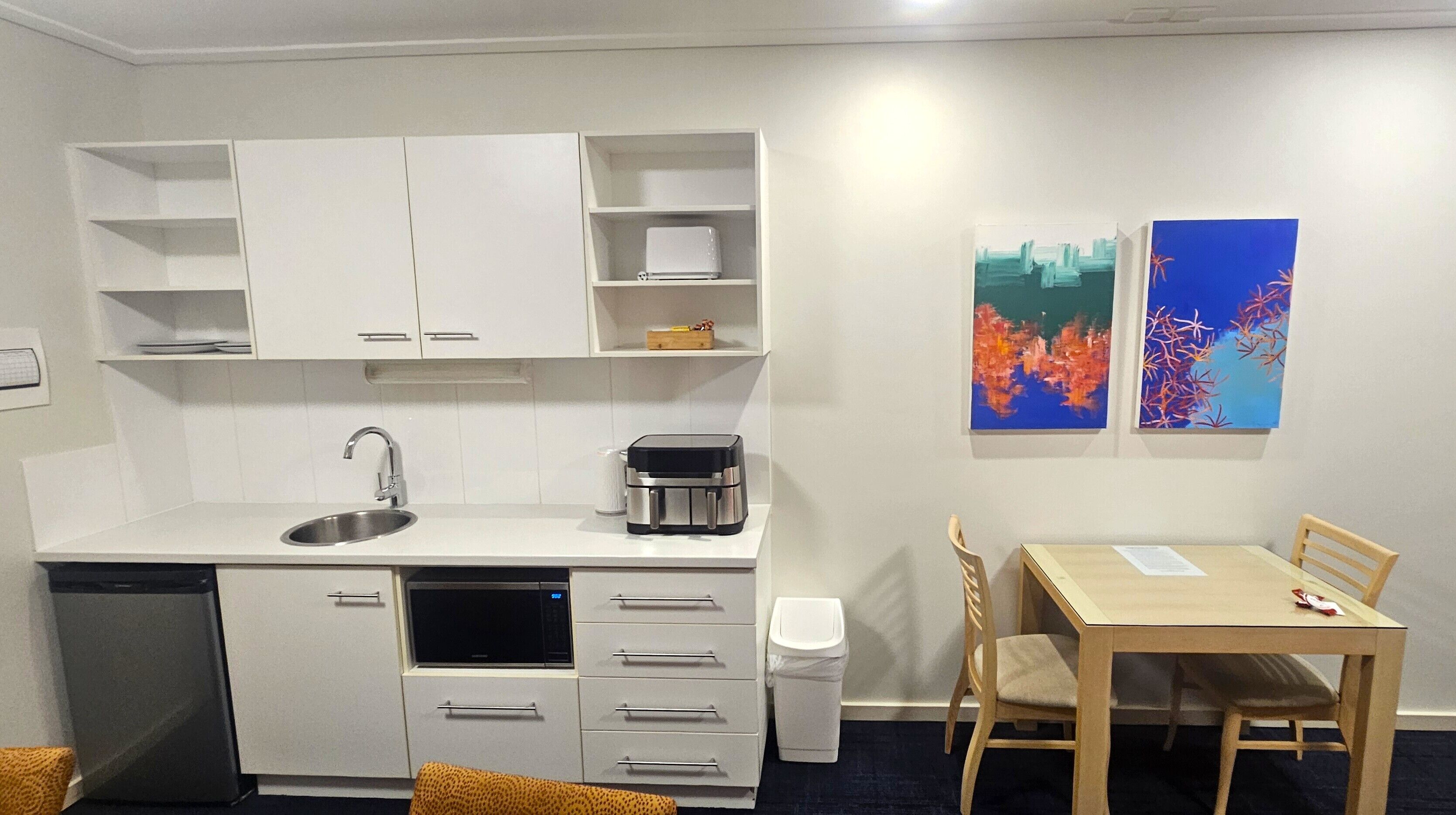 A compact kitchen with a dishwasher, microwave, and coffee maker. There is a dining table with two chairs and a trash bin. Two abstract paintings are on the wall.