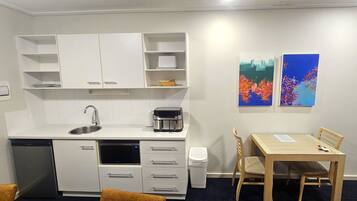 A compact kitchen with a dishwasher, microwave, and coffee maker. There is a dining table with two chairs and a trash bin. Two abstract paintings are on the wall.