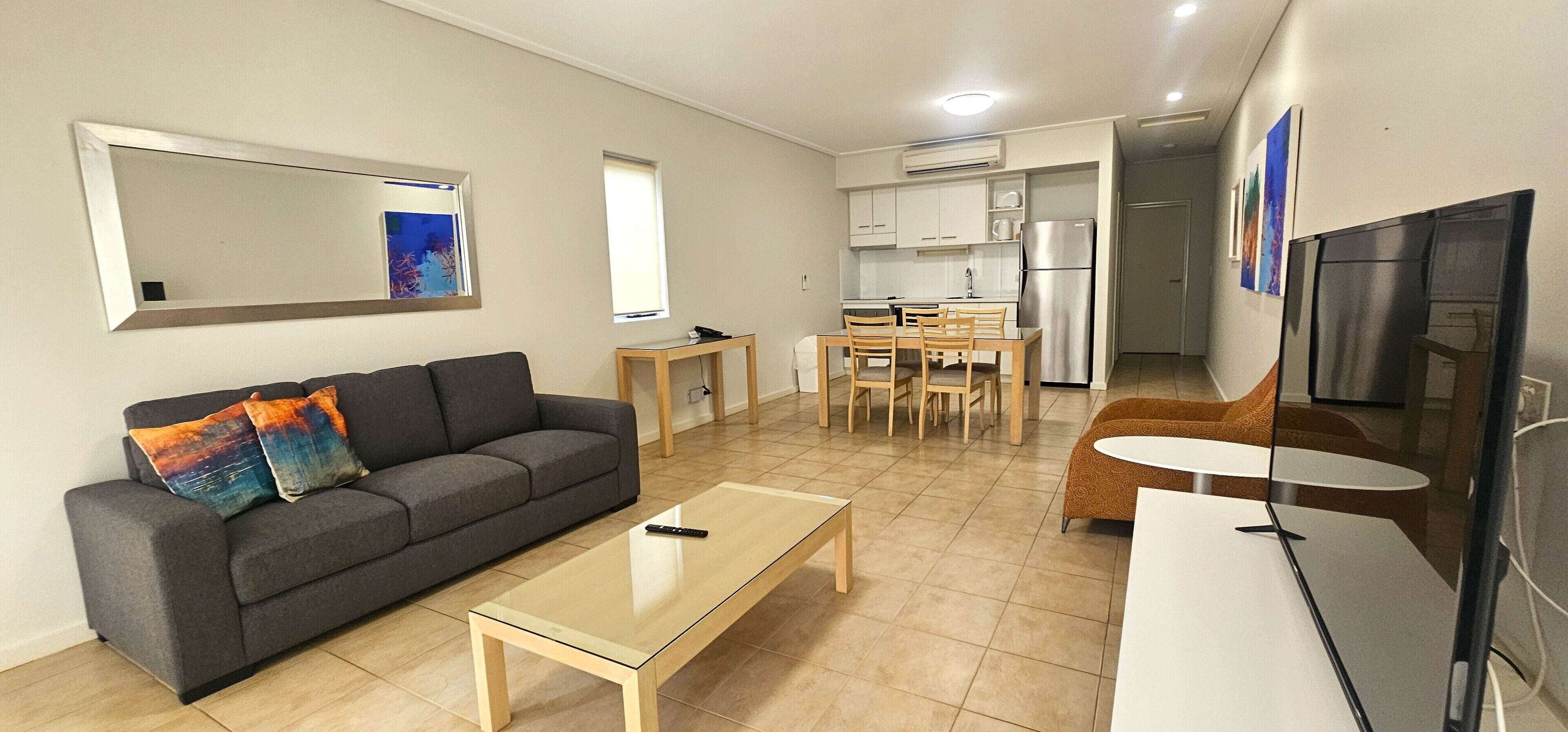 A modern living room with a grey sofa, a coffee table, and a TV. There is a dining area with a table and chairs. The kitchen is visible with a fridge and cabinets.
