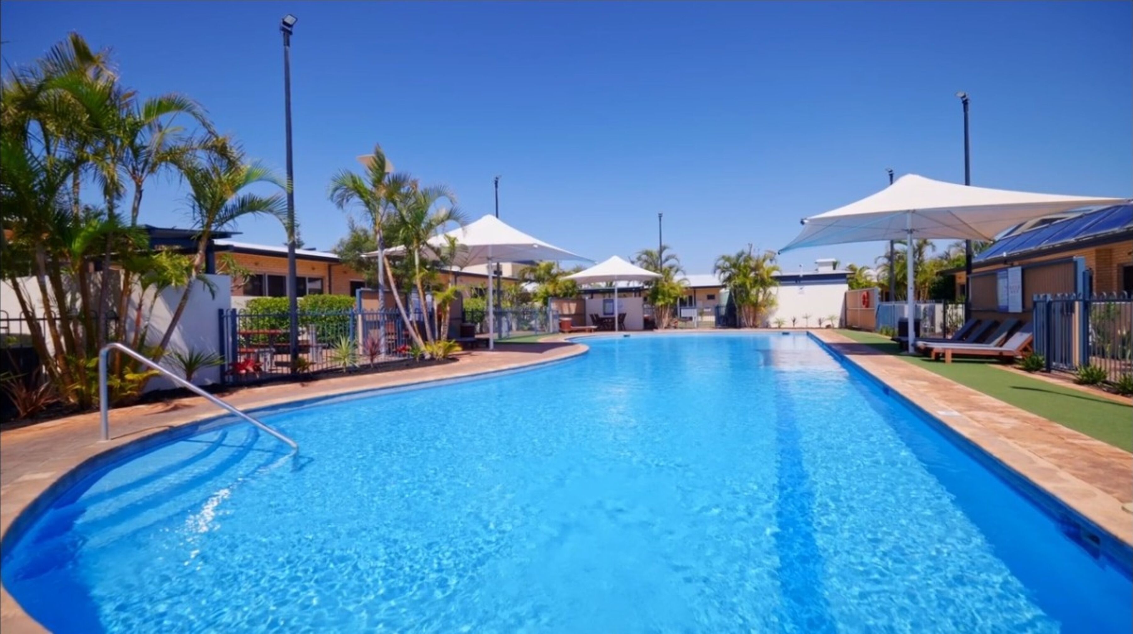 A swimming pool with clear blue water, surrounded by palm trees and umbrellas, and adjacent to a building with a flat roof.