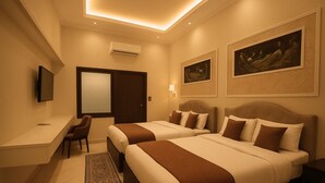 A hotel room with two beds, a desk, a chair, a TV, and artwork on the walls. - Hotel Om Vilas (Mathura)