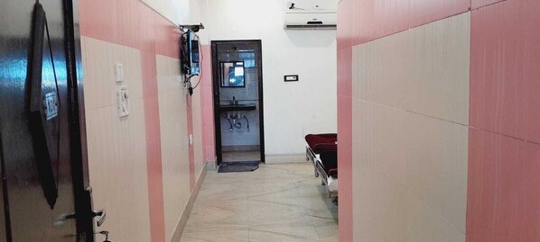 A narrow hallway with pink and white walls, a door at the end, and a bed with a red cover in the background. - BABA SITARAM ASHRAM (Haridwar)