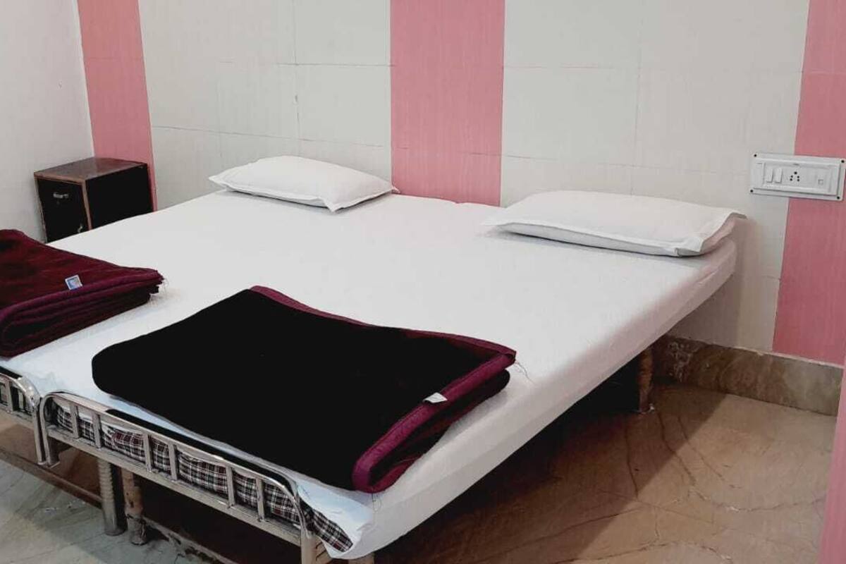 A single-bed room with a metal frame, a white mattress, two pillows, a folded red blanket, and a small black speaker.