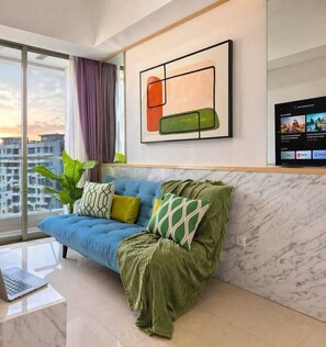 A modern living room with a blue sofa, green throw, and a large window with a city view. - Alona By SanLiving - 2BR - HubLife Mall (Jakarta)
