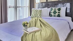A hotel room with a large bed, a bedside table with a plant and a book, a green blanket, and a large mirror. - Alona By SanLiving - 2BR - HubLife Mall (Jakarta)