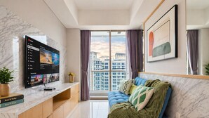 A modern living room with a large TV, a sofa, a coffee table, and a view of the cityscape through large windows. - Alona By SanLiving - 2BR - HubLife Mall (Jakarta)