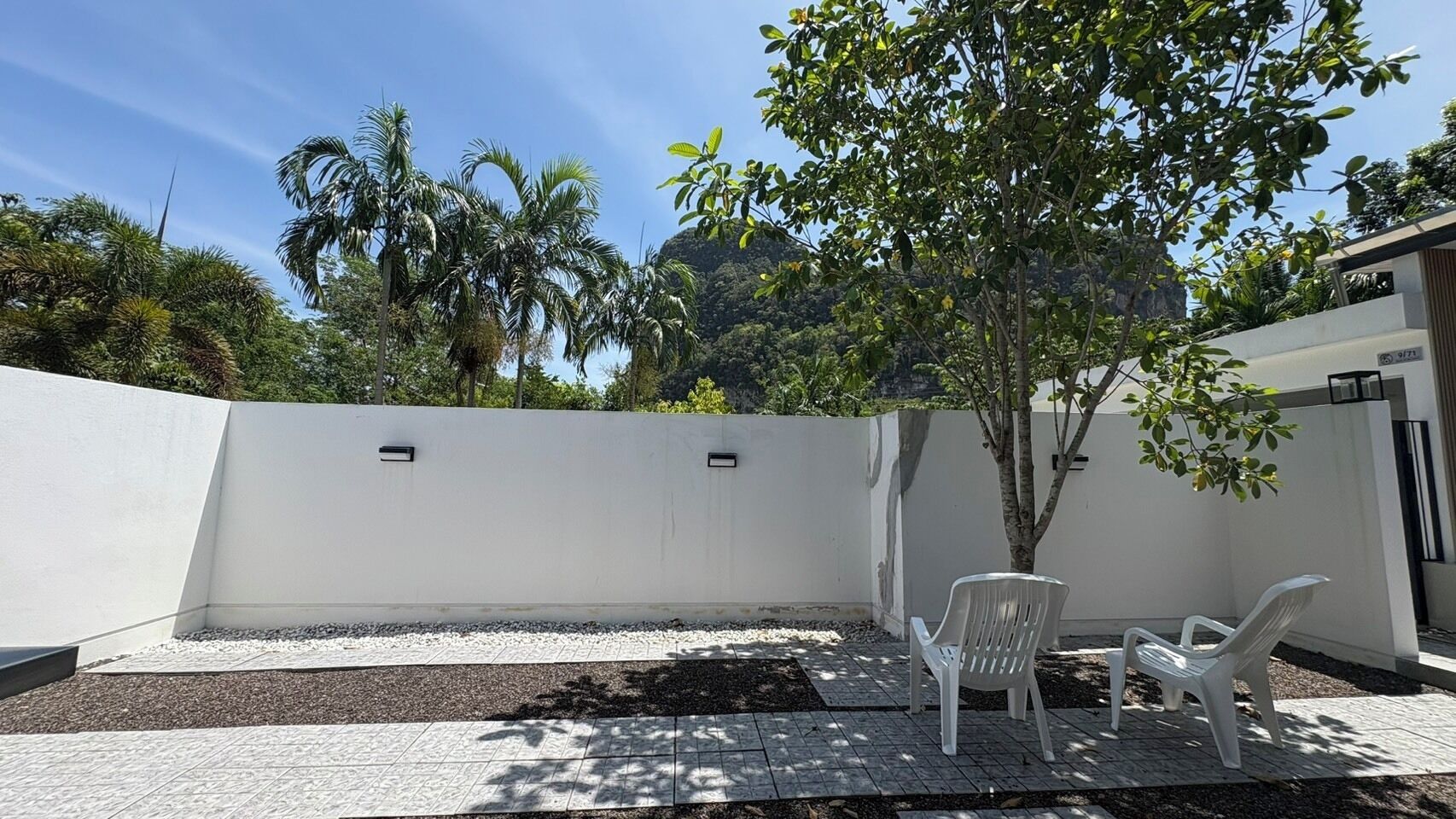 Un patio con una pared blanca, un árbol pequeño y dos sillas de plástico blancas.