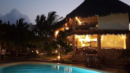 A poolside area with a thatched-roof structure, palm trees, and a well-lit outdoor space.