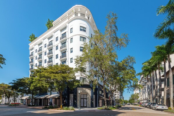 Ein mehrstöckiges Hotel mit Balkonen, einem überdachten Eingang und Blick auf die Straße mit parkenden Autos und Bäumen.