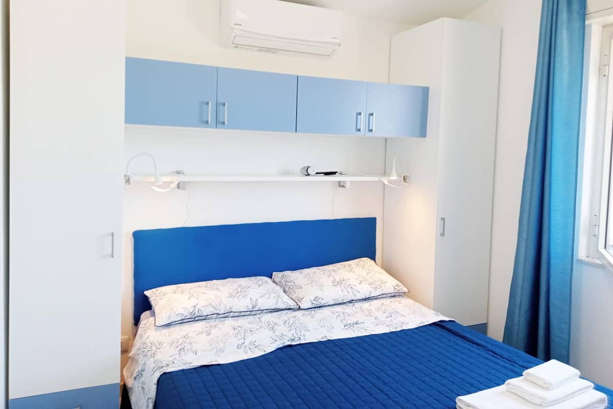 A bedroom with a blue bedspread, white floral pillows, a white air conditioner, and blue curtains.