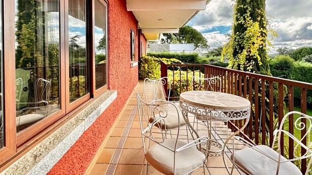 A balcony with a round table and chairs, a red textured wall, and a view of greenery and trees. - Apartment in Galapagar With Pool and Garden (Galapagar)