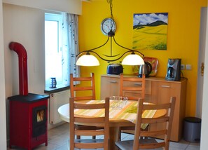 Un coin repas avec un poêle rouge, des chaises en bois, une table recouverte d’une nappe à rayures, une horloge, un tableau et des appareils de cuisine.
