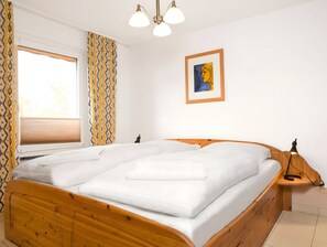 Un lit bien fait, avec du linge de lit blanc, dans une chambre dotée d’une tête de lit en bois, d’une fenêtre ornée de rideaux à motifs, d’un tableau abstrait encadré accroché au mur et d’une table de chevet avec une lampe.