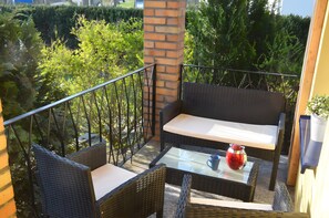 Un balcon agrémenté de meubles en osier, d’une table en verre et d’un vase rouge.