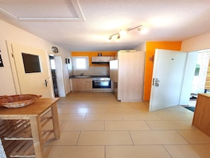 A compact kitchen with a sink, stove, and refrigerator, a dining table with a basket, and a window with blinds. - Feriendomizile auf der Insel Poel (Insel Poel)