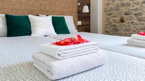 Una cama bien tendida con toallas blancas dobladas y un cojín decorativo con texto, sobre un edredón estampado y una pared de piedra.