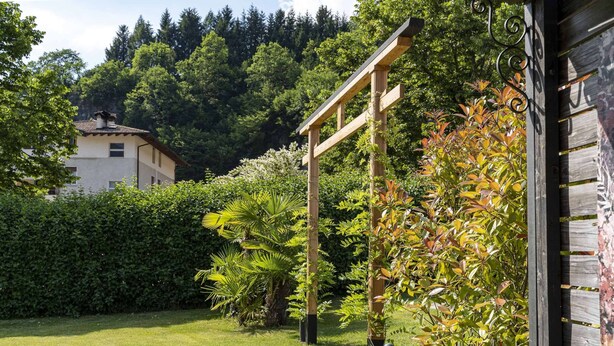 Un jardín con un pérgola de madera, abundante vegetación y un edificio al fondo.