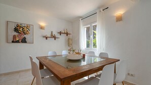 Un comedor con una mesa de vidrio, sillas blancas, un banco de madera, un cuadro, estanterías y una ventana con cortinas.