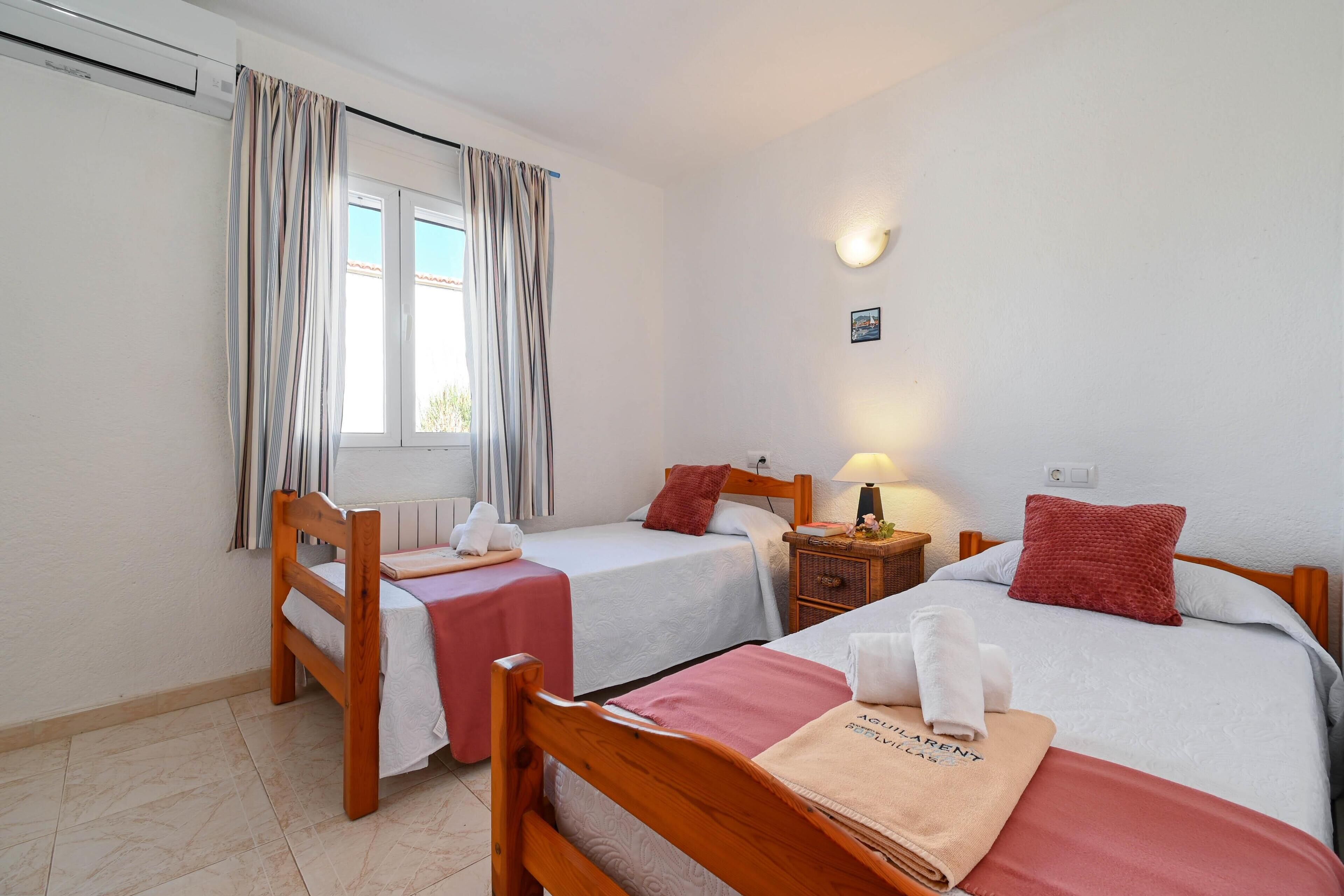 A hotel room with two single beds, a wooden chair, a small table, a nightstand with a lamp, and a window with curtains.