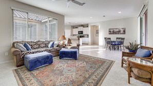Een ruime woonkamer met een grote bank, voetenbankjes en een vloerkleed met een patroon. Er is een plafondventilator, een eethoek met stoelen en een keuken op de achtergrond.