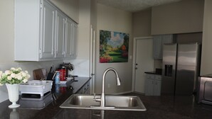 A kitchen with a stainless steel sink, white cabinets, a painting on the wall, and a refrigerator. - Peaceful Retreat  Near NASA & Kemah 3BR/2BA (Houston)