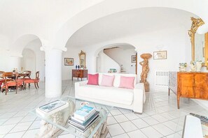 A spacious living room with a white sofa, a glass coffee table, and a dining area with wooden chairs and a table. - Villa Uma: A bright and sunny two-story villa which faces the sun and the sea, with Free WI-FI. (Praiano)
