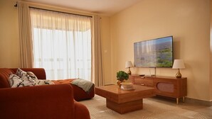 A modern hotel room with a large window, a flat-screen TV, a wooden coffee table, a sofa, and a side table with a lamp. - 2-bedroom apartment with Lake views and a heated pool in Nakuru Town (Nakuru)
