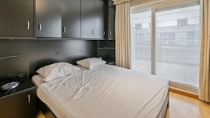 A modern bedroom with a large bed, built-in dark wooden wardrobe, and a large window with a view of buildings. - Cozy four bedroom apartment by the harbor (Blankenberge)
