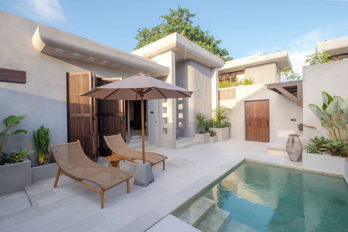A modern outdoor pool area with lounge chairs and a large umbrella, surrounded by minimalist architecture and potted plants.