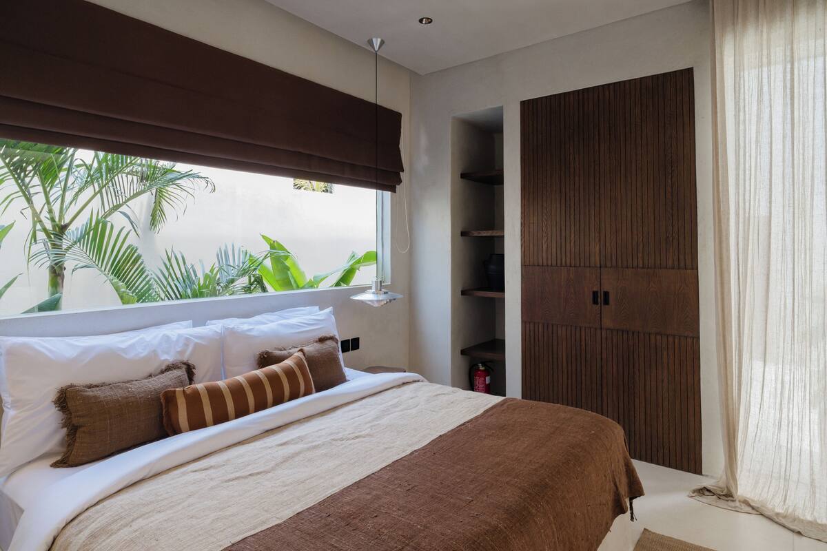 A bedroom with a bed, a window with a view of greenery, a wooden wardrobe, and a bedside table with a lamp.