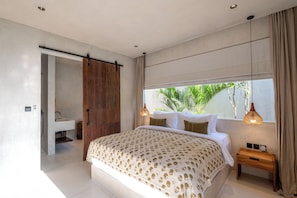 A bedroom with a bed, a nightstand, a window with a view of greenery, a sliding door to a bathroom, and a pendant light. - Heritage Villas (Pecatu)