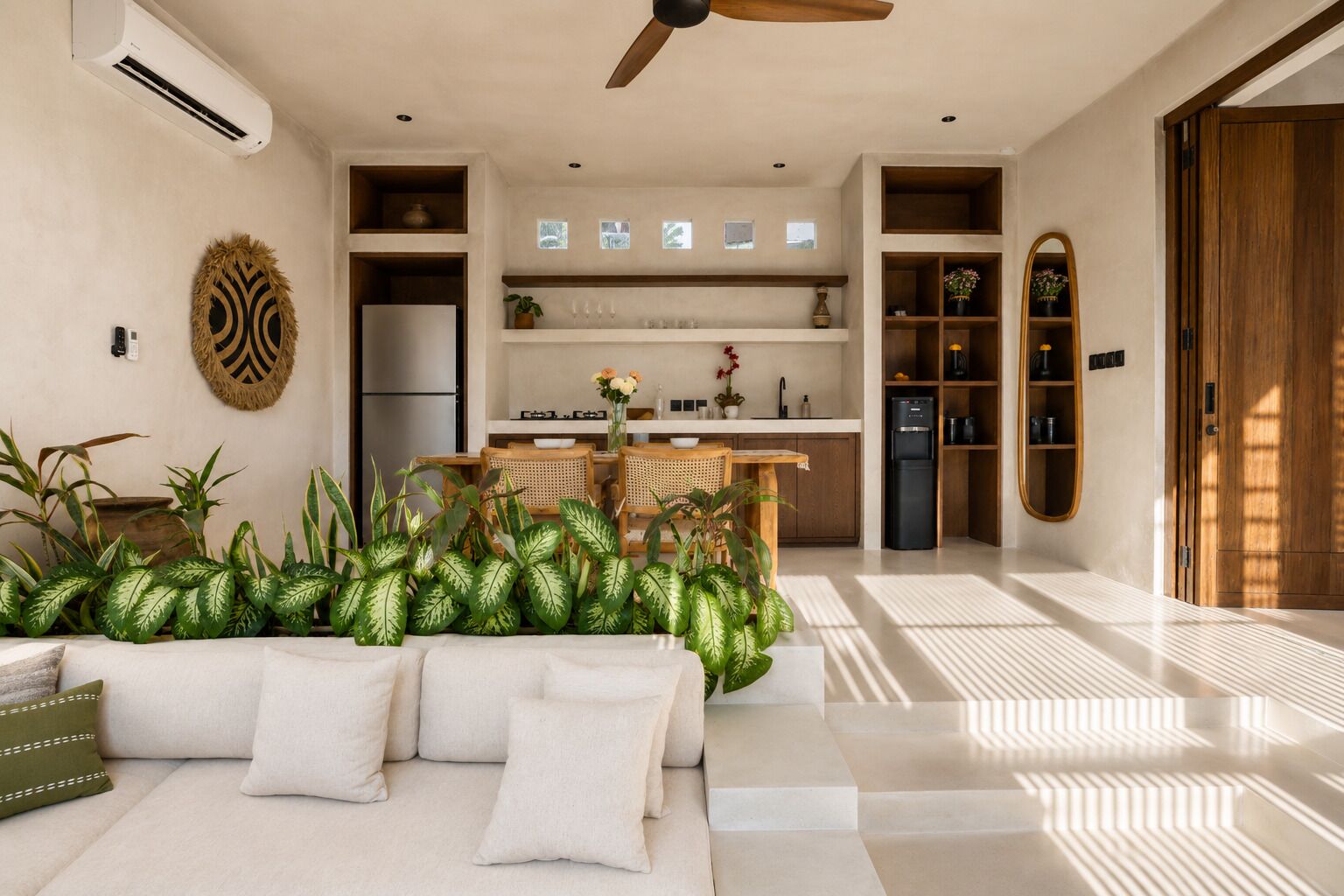 A modern living room with a sofa, coffee table, and plants. There is a kitchen area with a refrigerator, stove, and shelves.