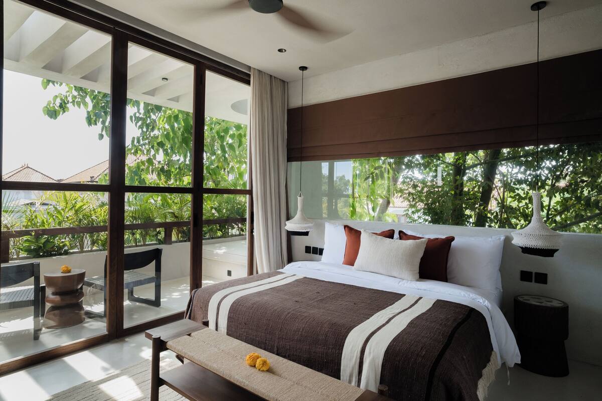 A modern bedroom with a large bed, a bench, and a view of greenery outside.