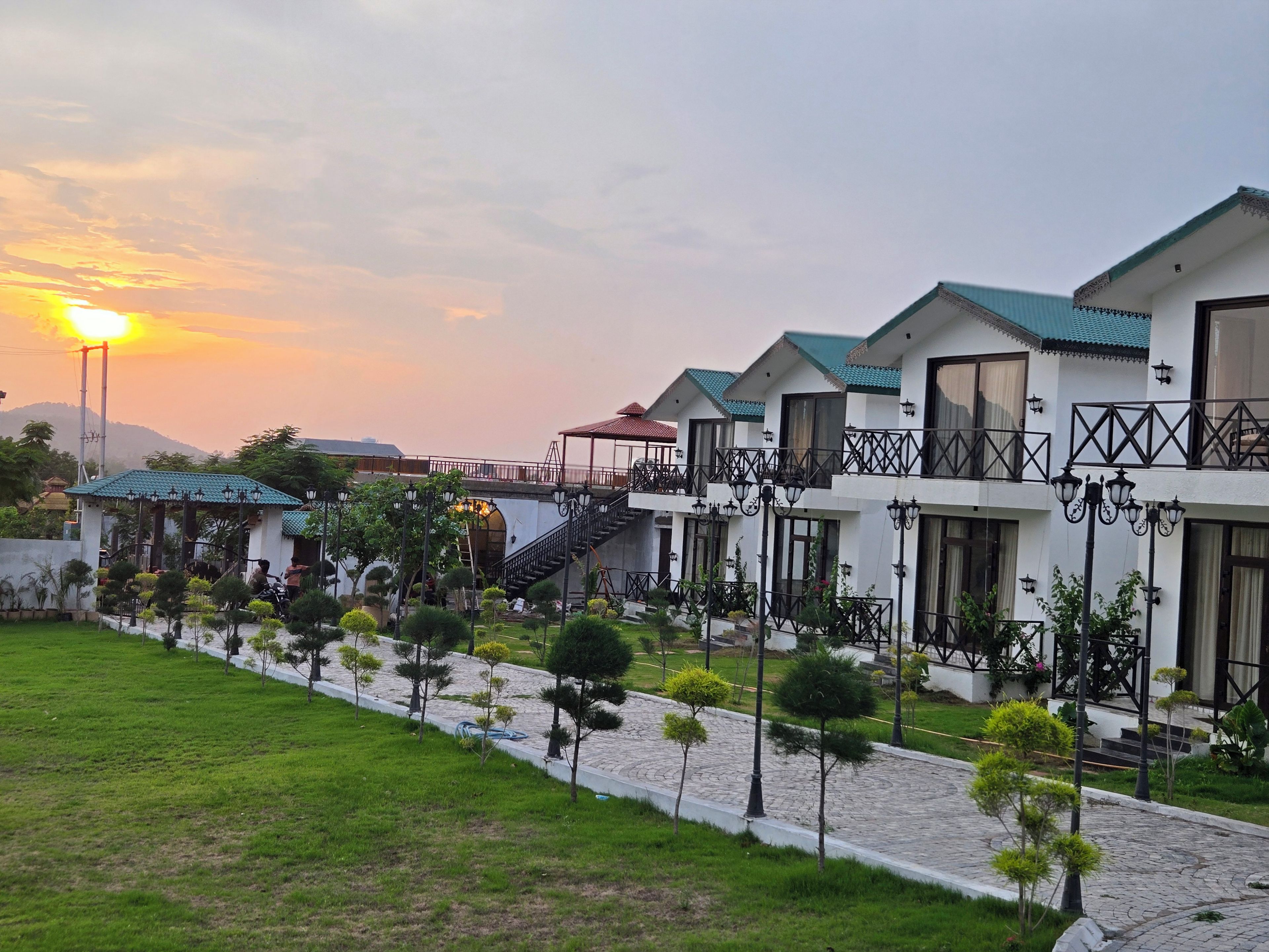 A row of modern buildings with balconies, a paved walkway, and a sunset in the background.