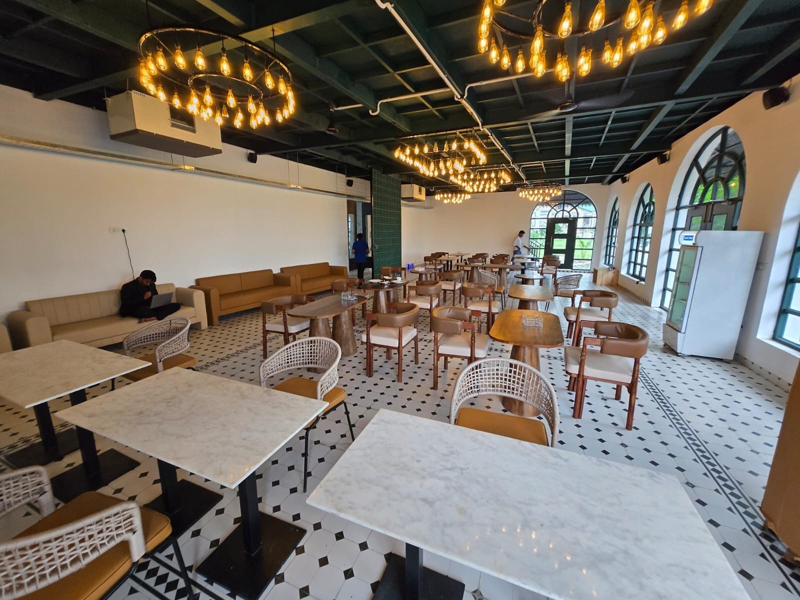 A spacious cafe with multiple seating areas, including tables and chairs, and a high ceiling with exposed beams and hanging light fixtures.