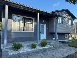 A modern two-story house with a grey exterior, a white door, and a small front garden with white pebbles and green shrubs. - Warm and friendly Canadian home 20 minutes from Montreal (Mascouche)