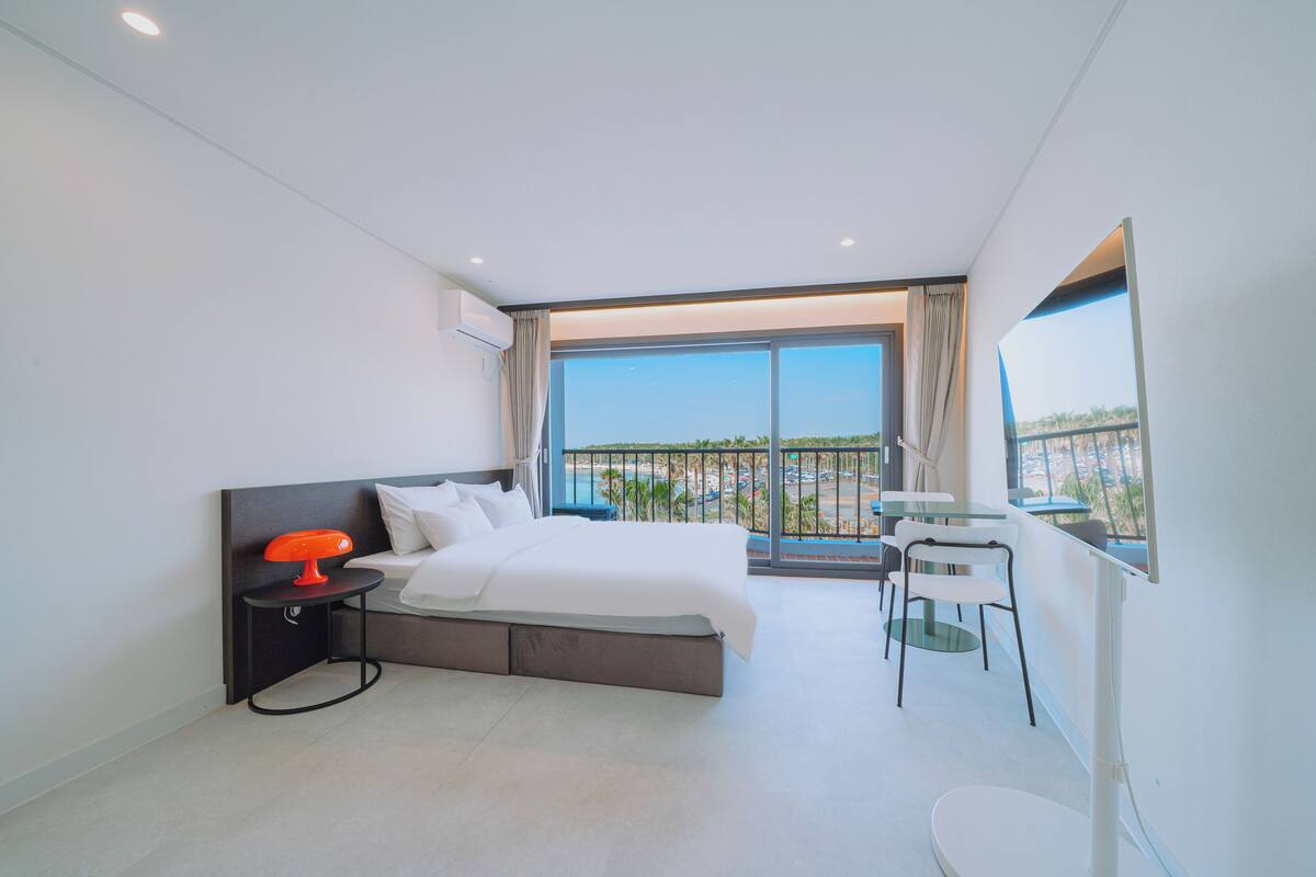 Ein modernes Hotelzimmer mit einem großen Bett, einem kleinen Tisch, einem Sessel und Blick ins Freie.
