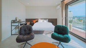 A modern hotel room with a large bed, two chairs, a small table, and a balcony with a city view. - Geumneung inn (Jeju City)