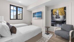 Habitación de hotel moderna con un ventanal grande, una cama con ropa de cama blanca, un sillón gris y un televisor montado en la pared.