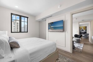A modern bedroom with a large bed, a window with a city view, a painting of a beach, and a hallway leading to a dining area.