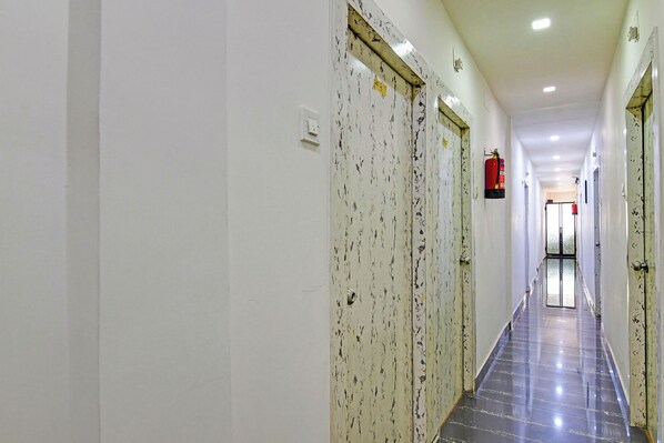 Un pasillo largo y estrecho con paredes y puertas blancas, un extintor fijado a la pared y suelo de baldosas.