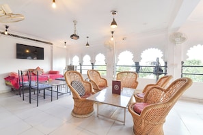A room with a dining table, wicker chairs, a flat-screen TV, and large windows with decorative arches. - Fabhotel Boutique Stays Lake Pichola (Udaipur)