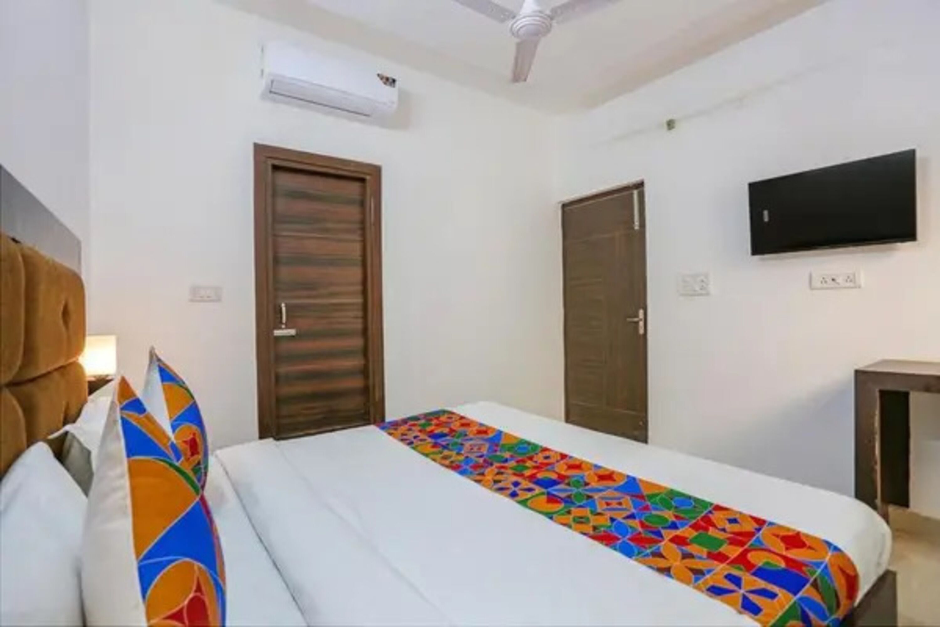 A hotel room with a bed, a ceiling fan, an air conditioning unit, a TV mounted on the wall, and two doors.