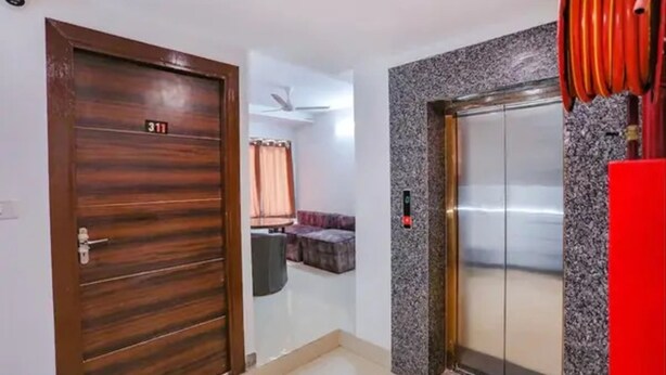 Un pasillo con una puerta de madera con la etiqueta “311”, un ascensor con cable rojo y una zona de estar con ventilador de techo.