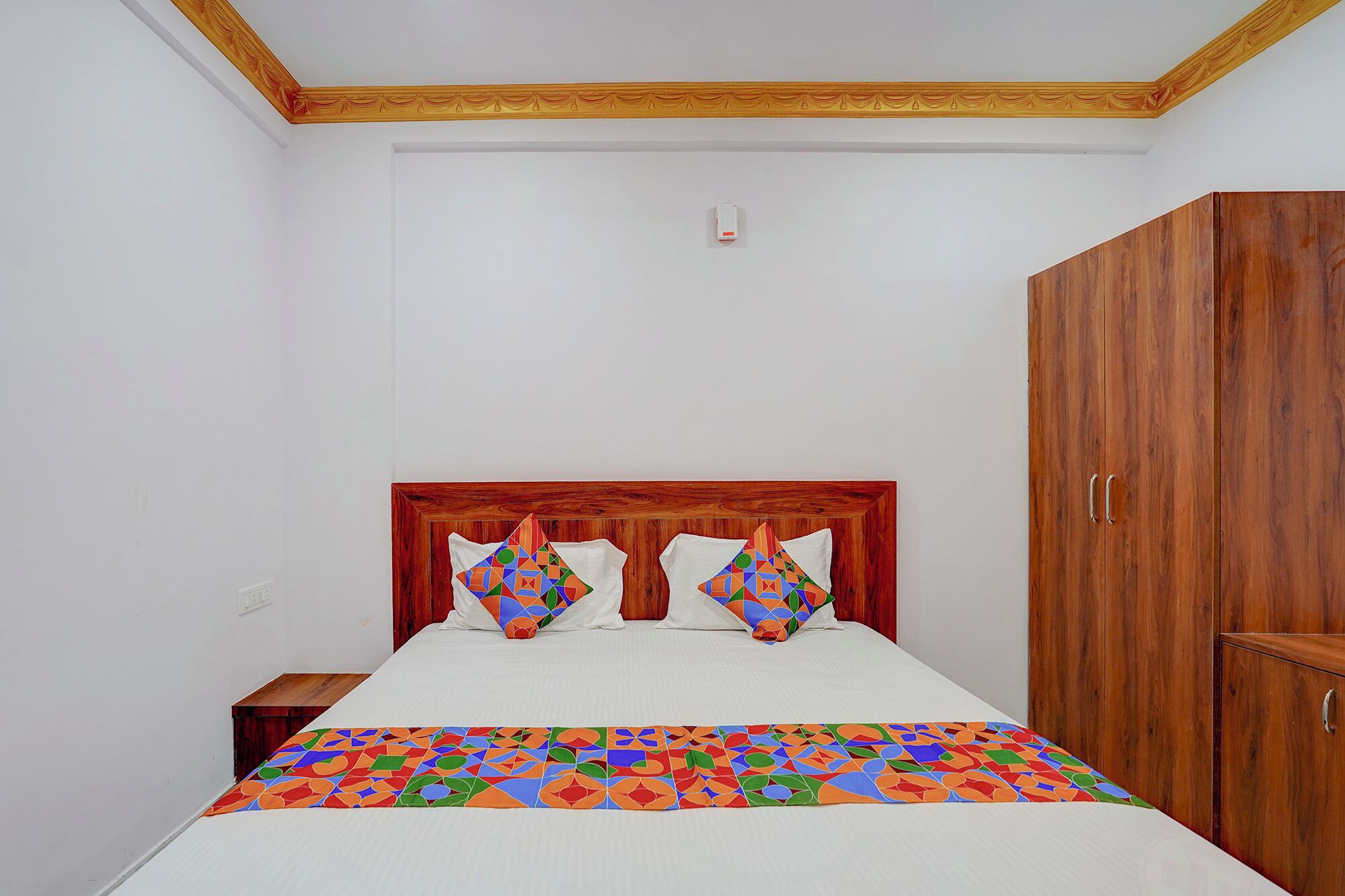 Ein Schlafzimmer mit einem hölzernen Kopfteil, einem Bett mit weißer Bettwäsche, zwei Kissen mit bunten geometrischen Mustern, einem Holzschrank und einem kleinen Nachttisch.