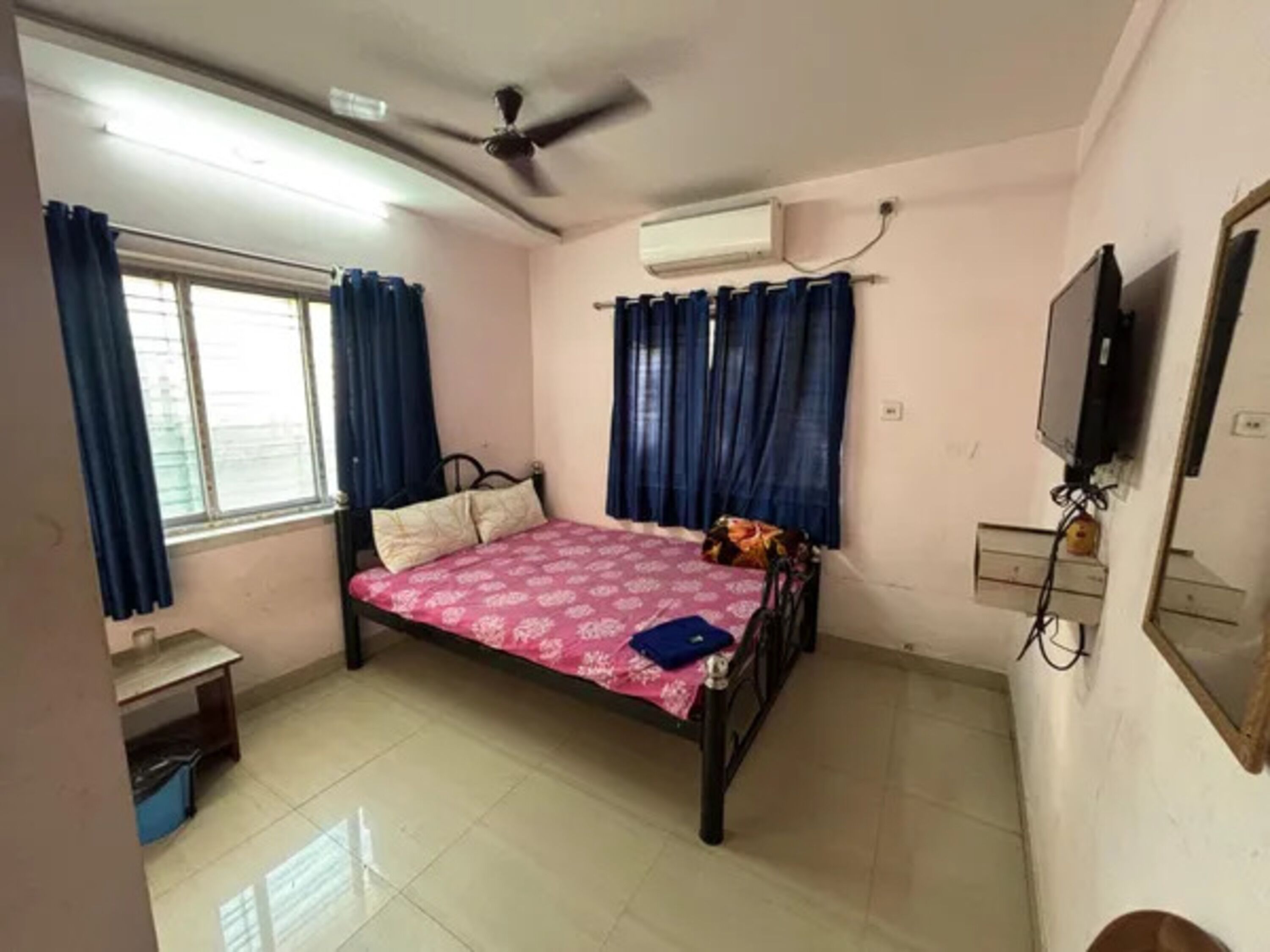 A bedroom with a bed, a ceiling fan, an air conditioner, a TV, and a window with curtains.
