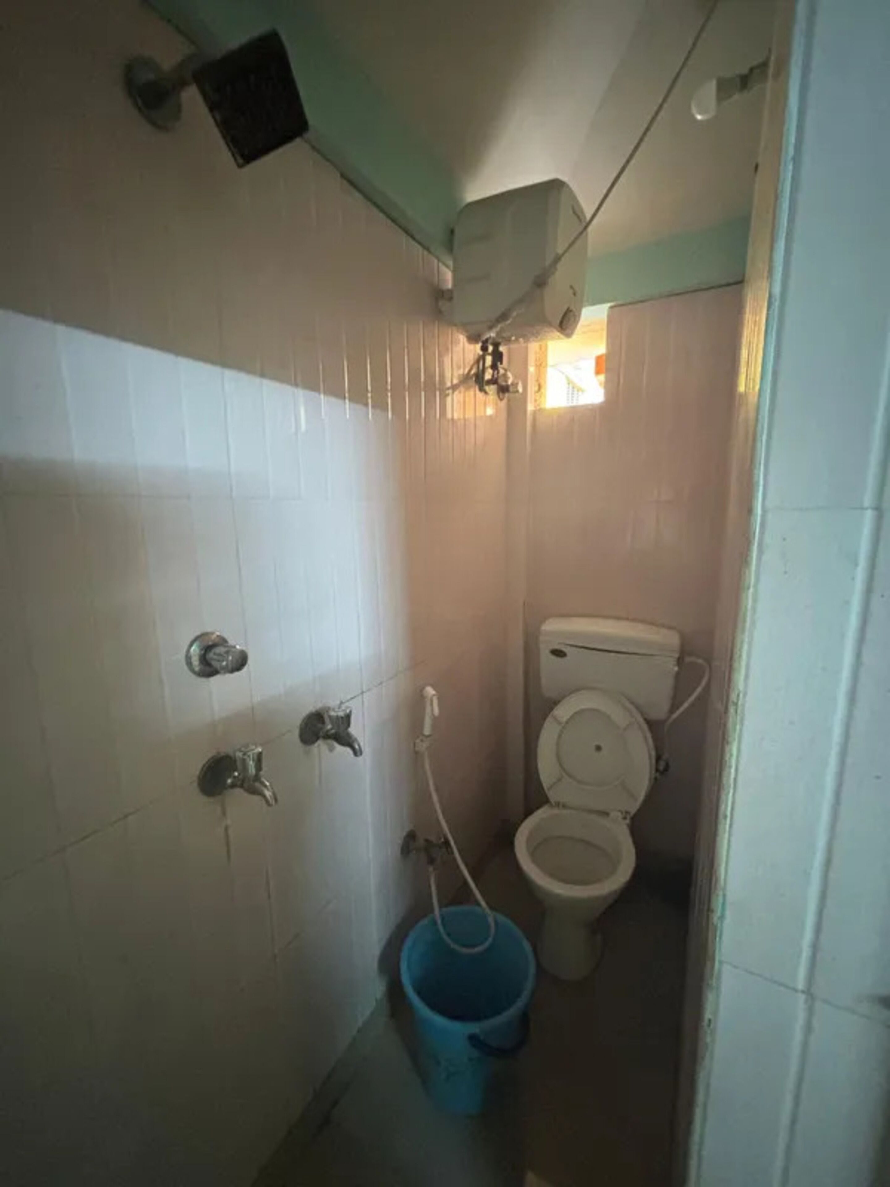 Un baño pequeño y estrecho con paredes de azulejos blancos, un inodoro, un cabezal de ducha y un cubo azul.